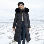 Woman wearing black longline puffer coat with black fur hood and jeans, standing on snowy terrain