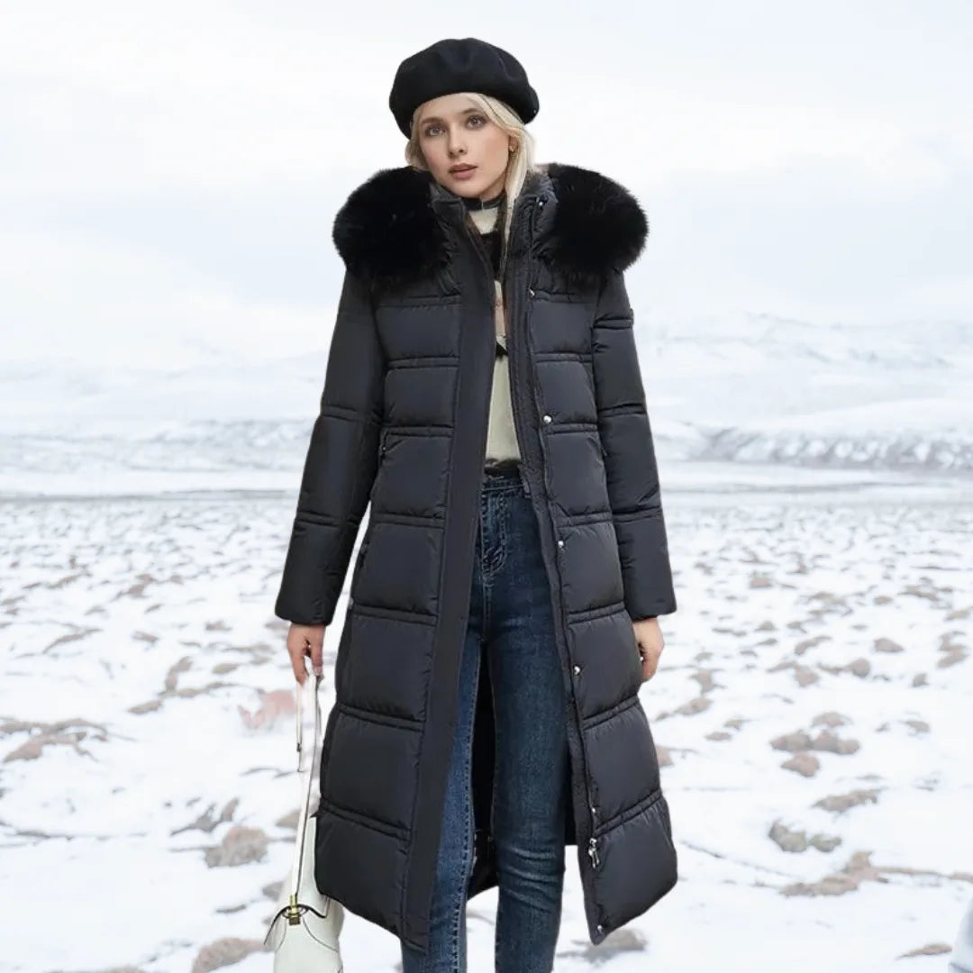 Woman wearing black longline puffer coat with black fur hood and jeans, standing on snowy terrain