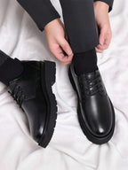 Overhead view of a hand adjusting a sock above a pair of black leather lace-up derby shoes on a white backdrop.