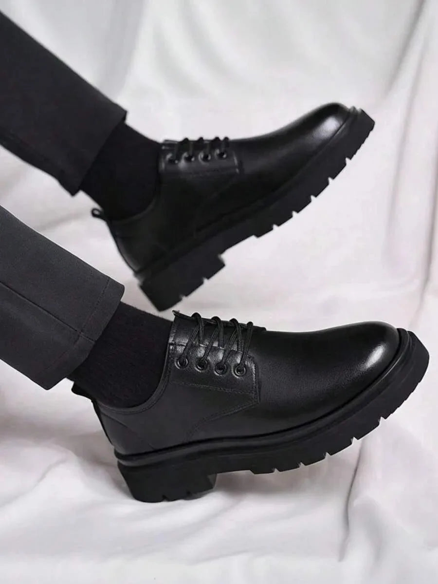 Close-up side profile of two black leather derby shoes worn with cropped trousers, showing the thick tread sole.