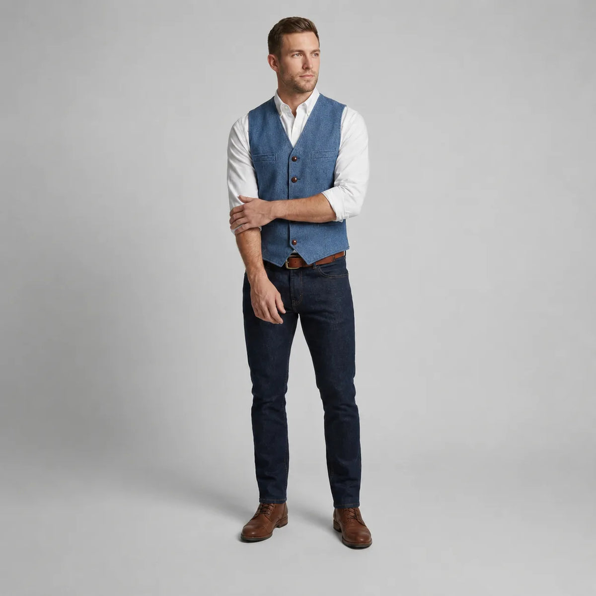 Full-length front view of a man in a blue waistcoat with crossed arms, paired with dark denim and brown boots