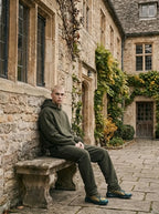 Man seated on a stone bench outside a countryside building wearing the olive green hoodie and jogger set