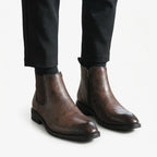 Close-up of feet wearing brown leather Chelsea boots with visible side zip, paired with black tailored trousers