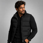 Man wearing a black quilted gilet open over a black polo neck jumper, hands in trouser pockets