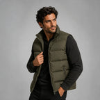 Man wearing an open army green quilted gilet over a black jumper, holding the collar with one hand