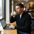 Man in a coffee shop drinking from a mug while wearing a black quarter-zip jumper over a white tee and jeans