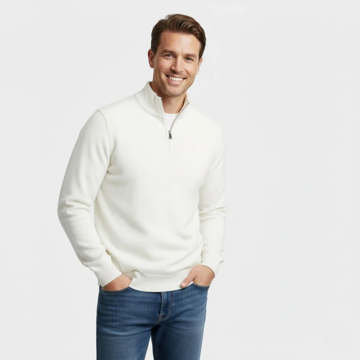 Man wearing an off-white quarter-zip jumper with a white tee underneath, hands in jean pockets, smiling
