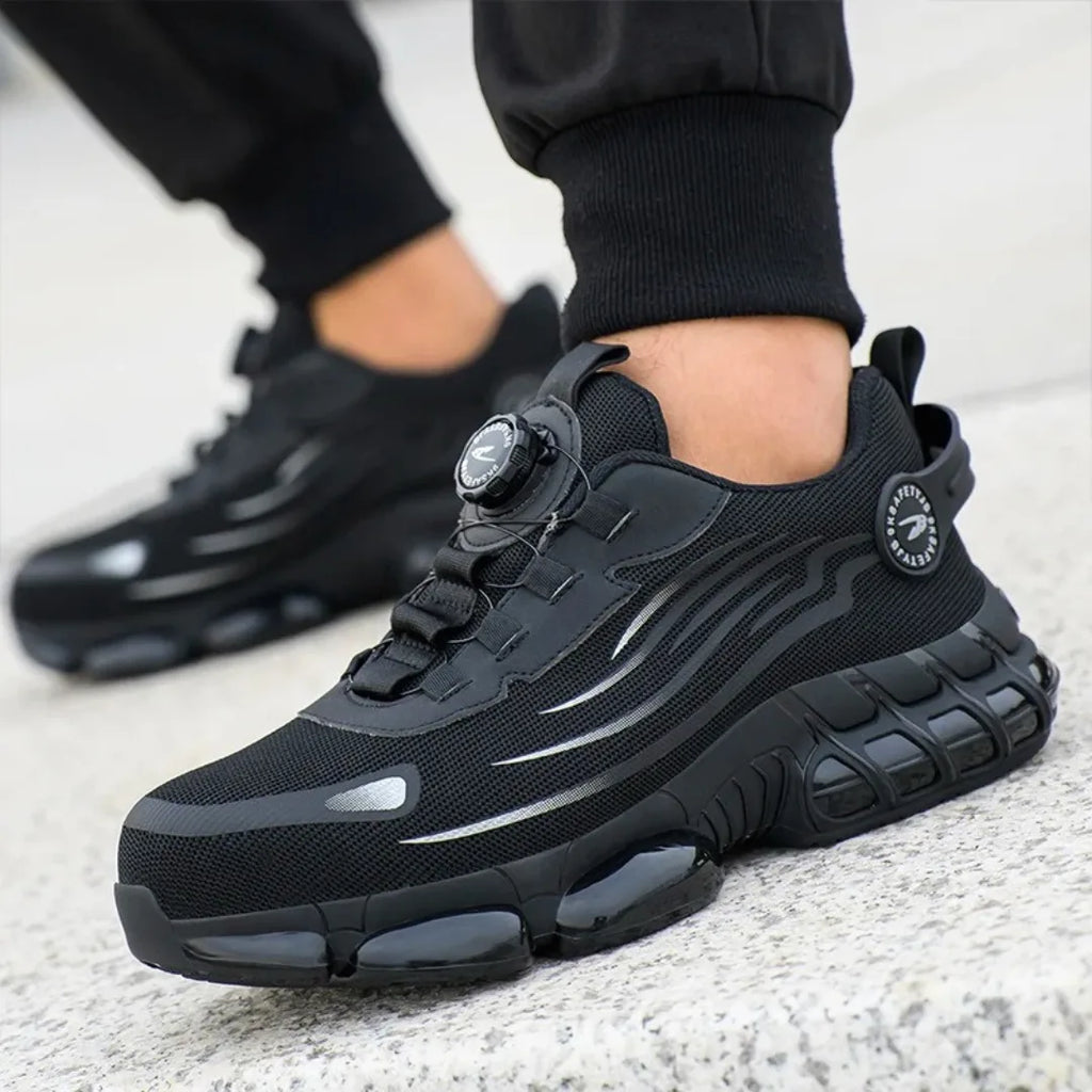 Close-up of a black safety trainer worn on foot, showing the rotary dial closure and reflective side detailing on concrete