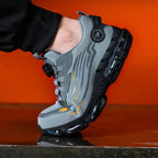 Close-up side profile of a grey safety trainer with black ridged sole worn on foot against an orange backdrop