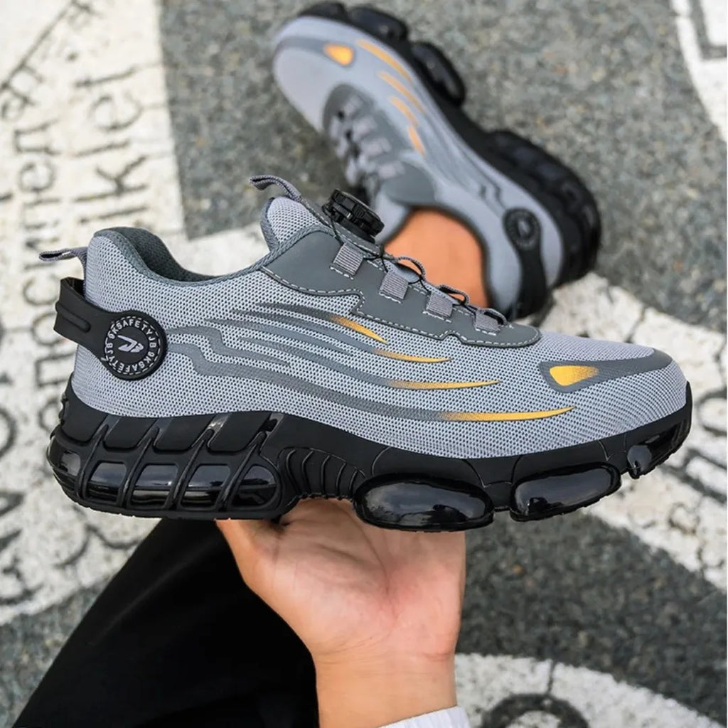 Grey mesh safety trainer with gold accent details held in hand, with its matching pair visible blurred in the background