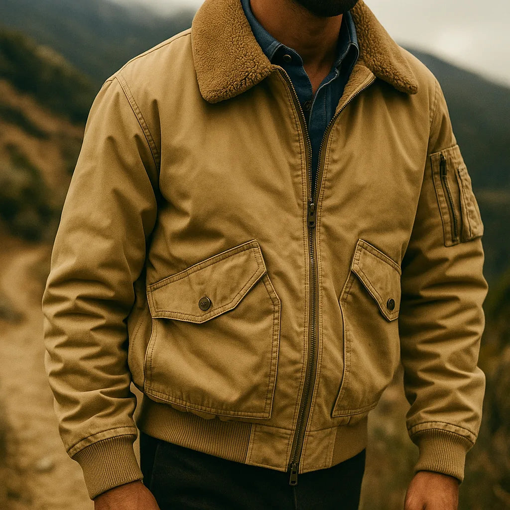 Tan bomber jacket with sherpa collar worn open, layered over a navy denim shirt, outdoor hillside setting