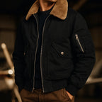 Black bomber jacket with tan shearling collar worn open over a dark jumper, sleeve zip pocket visible