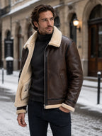 Man in brown leather aviator jacket with cream shearling collar, worn open over a black polo neck, standing on a snowy street.