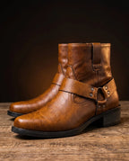 Side view of tan brown harness boots with buckle strap and studs, shown as a pair on a wooden surface