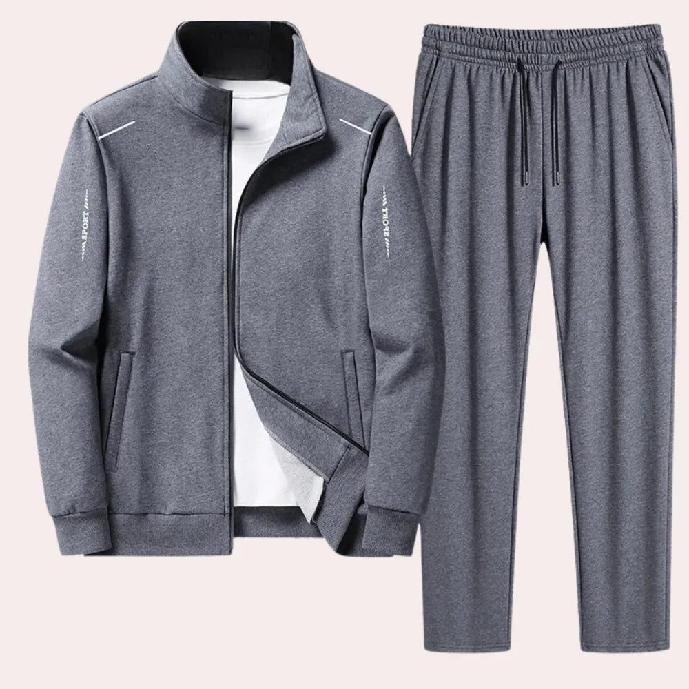 Grey zip-up tracksuit jacket with reflective shoulder trim worn over a white t-shirt, paired with matching grey joggers