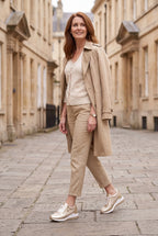 Woman in a beige trench coat and tailored trousers walking outdoors wearing gold metallic wedge trainers