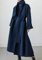 Front angled view of a navy polo neck maxi coat with wraparound scarf-style collar and tie waist, worn open over a black outfit
