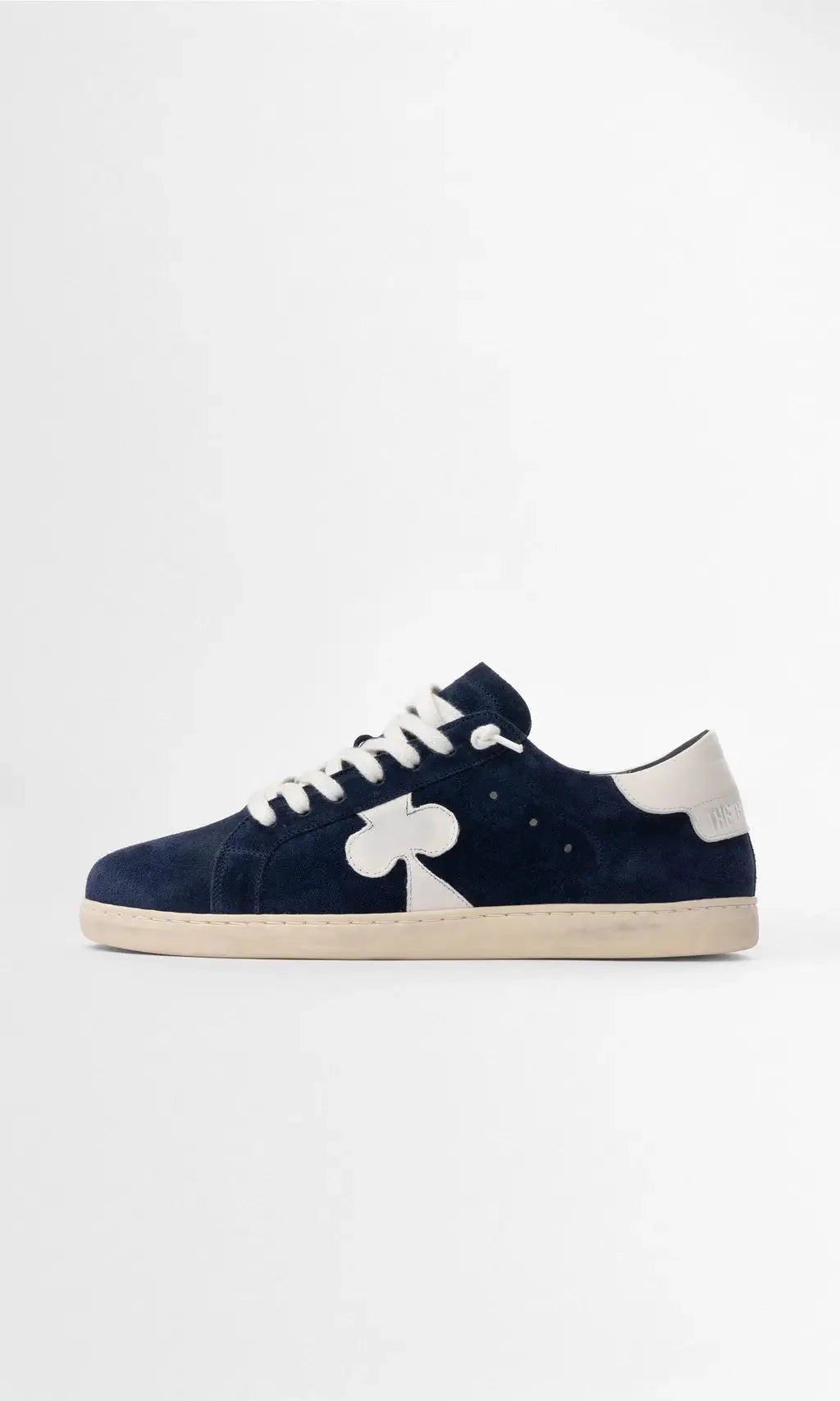 Side profile of navy suede trainer with white leather heel tab and clover-shaped side panel on a cream sole