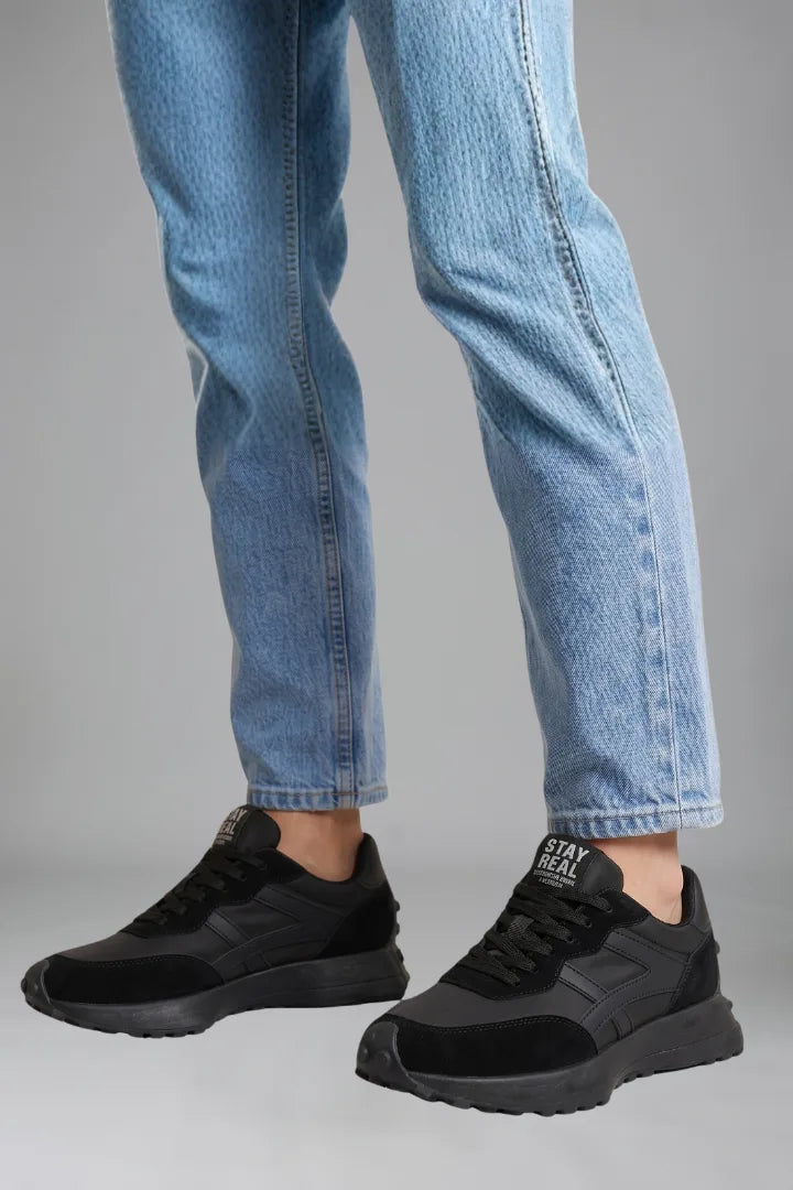 Side view of all-black trainers with tonal panels and matte finish worn under denim