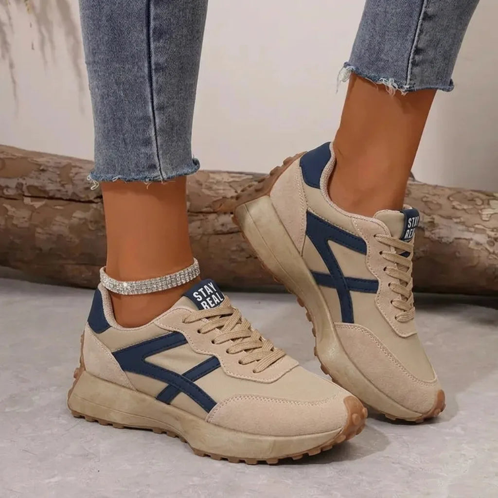 Beige suede trainer with navy panel detailing and lace-up front, worn on foot beside a wooden log