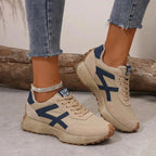 Beige suede trainer with navy panel detailing and lace-up front, worn on foot beside a wooden log