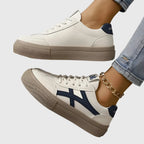 Pair of white trainers with navy heel tab and beige platform sole, one worn on foot with gold anklet