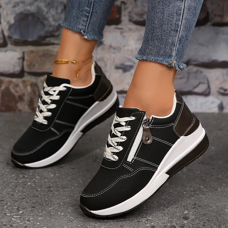 Pair of black lace-up trainers with bronze heel trim and white wedge sole shown against a stone wall