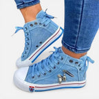 Side view of light blue denim high-top trainers with lace-up front and skull charm zip detail, worn with cropped jeans