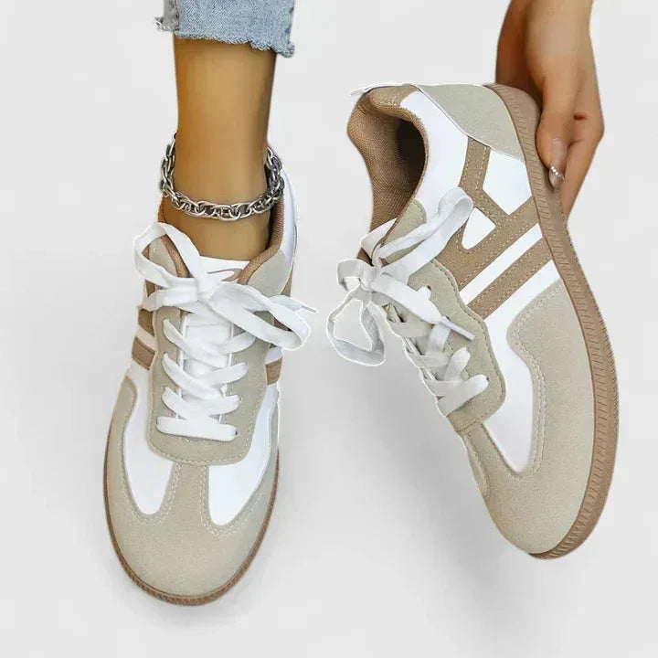 Top-down view of a matching pair of khaki suede and white leather trainers, one held by hand to show the padded collar