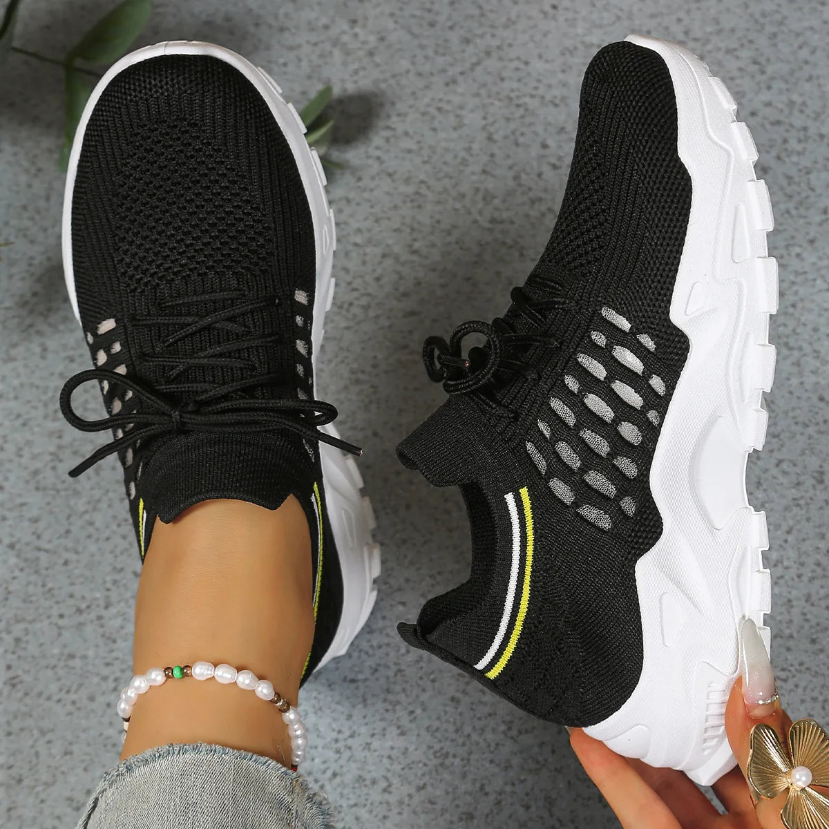 Overhead view of black knit trainers with white sole and yellow stripe detail, one shoe held by hand
