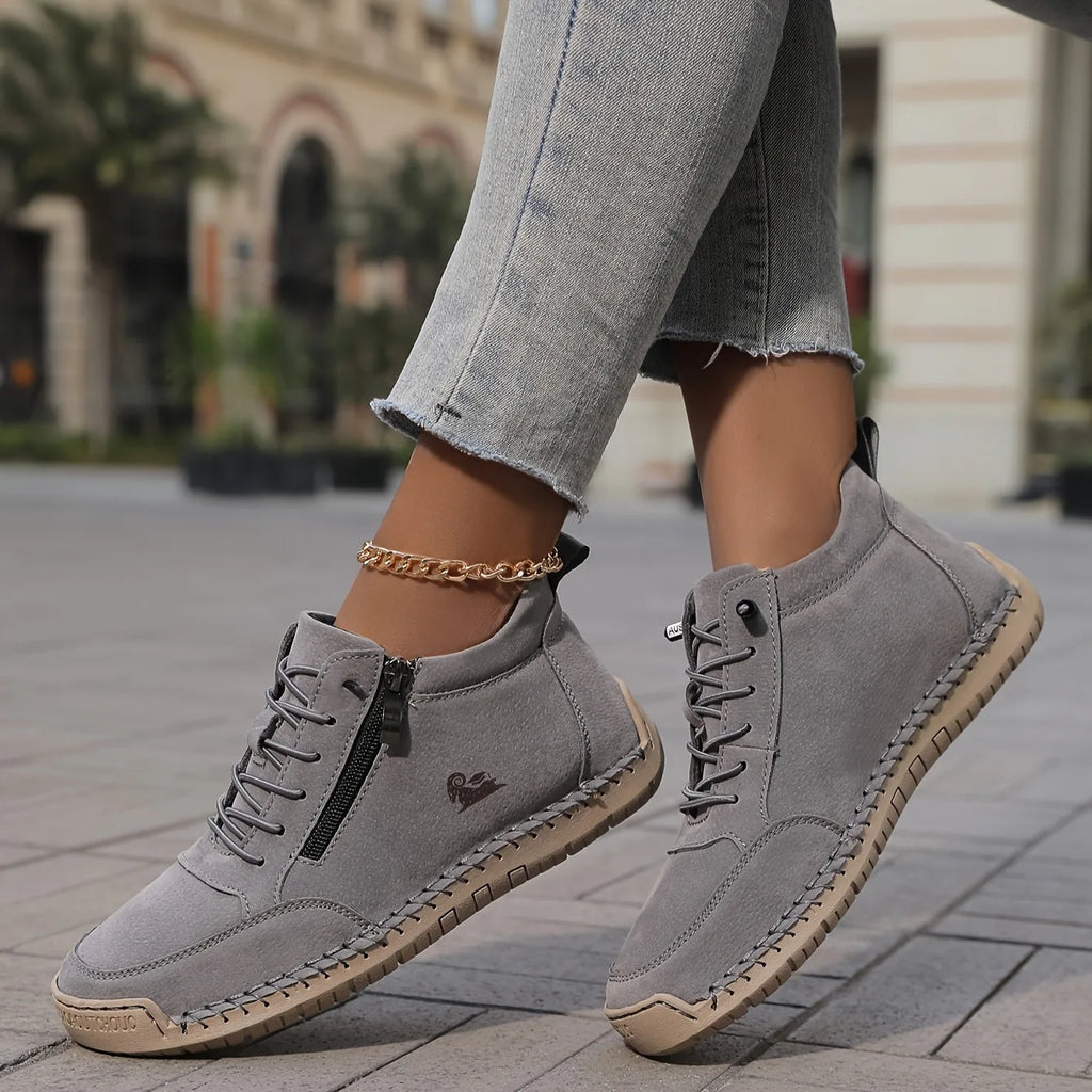 Pair of grey suede ankle sneakers with zip closure, lace-up front and tan textured sole on paving stones