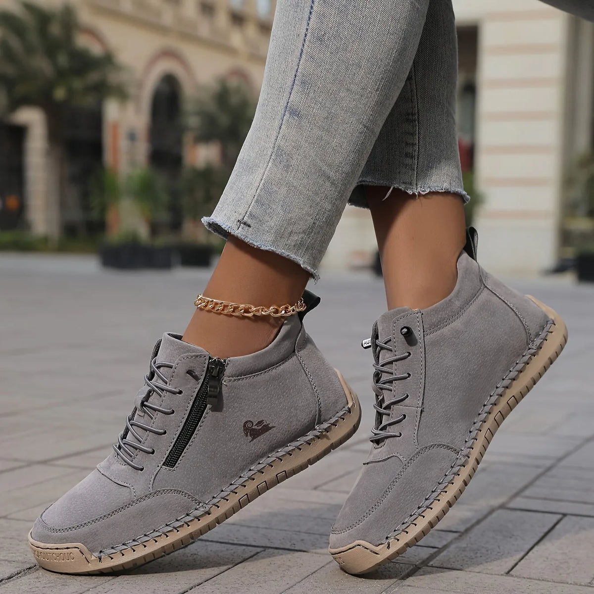 Pair of grey suede ankle sneakers with zip closure, lace-up front and tan textured sole on paving stones