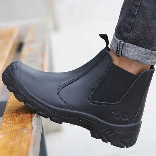 Side view of a black leather Chelsea boot worn with rolled-up jeans, showing the pull tab and reinforced toe cap