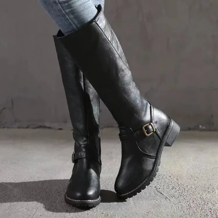 Side view of black leather knee-high boot with buckle strap detail, worn with jeans on concrete backdrop