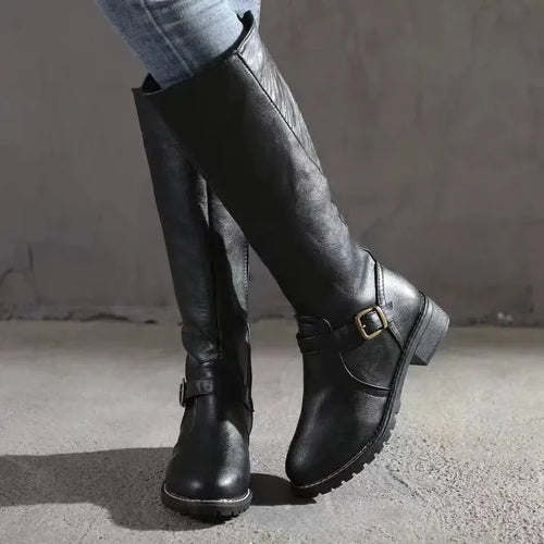 Side view of black leather knee-high boot with buckle strap detail, worn with jeans on concrete backdrop