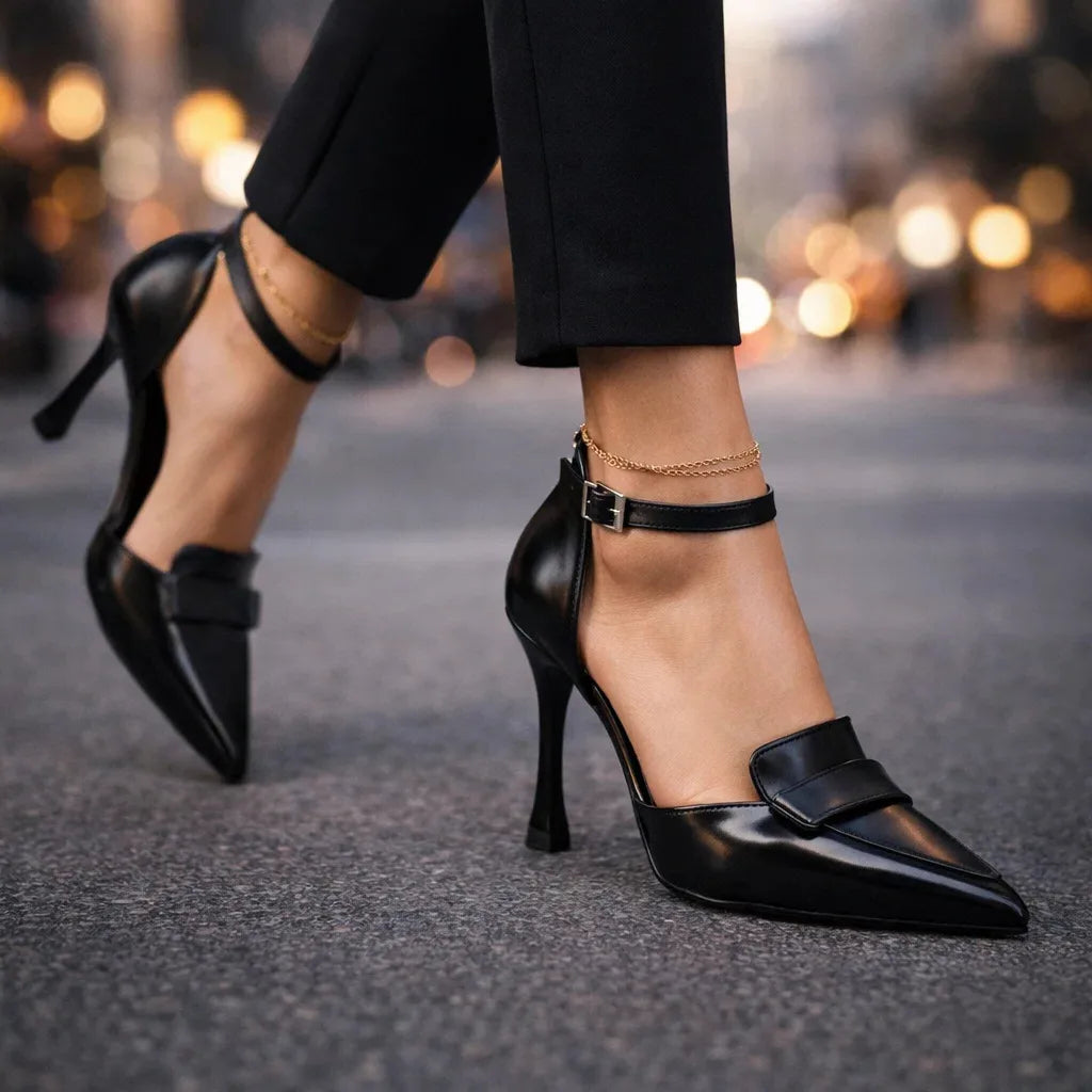 Walking shot of black pointed-toe heels with ankle strap, paired with black tailored trousers