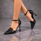 Close-up of black pointed-toe heel showing gold ankle chain layered over the buckled strap