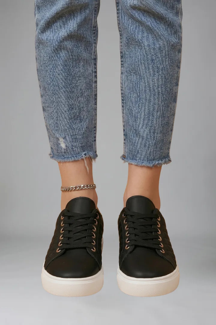 Close-up front view of black quilted trainers worn with cropped jeans and a silver anklet