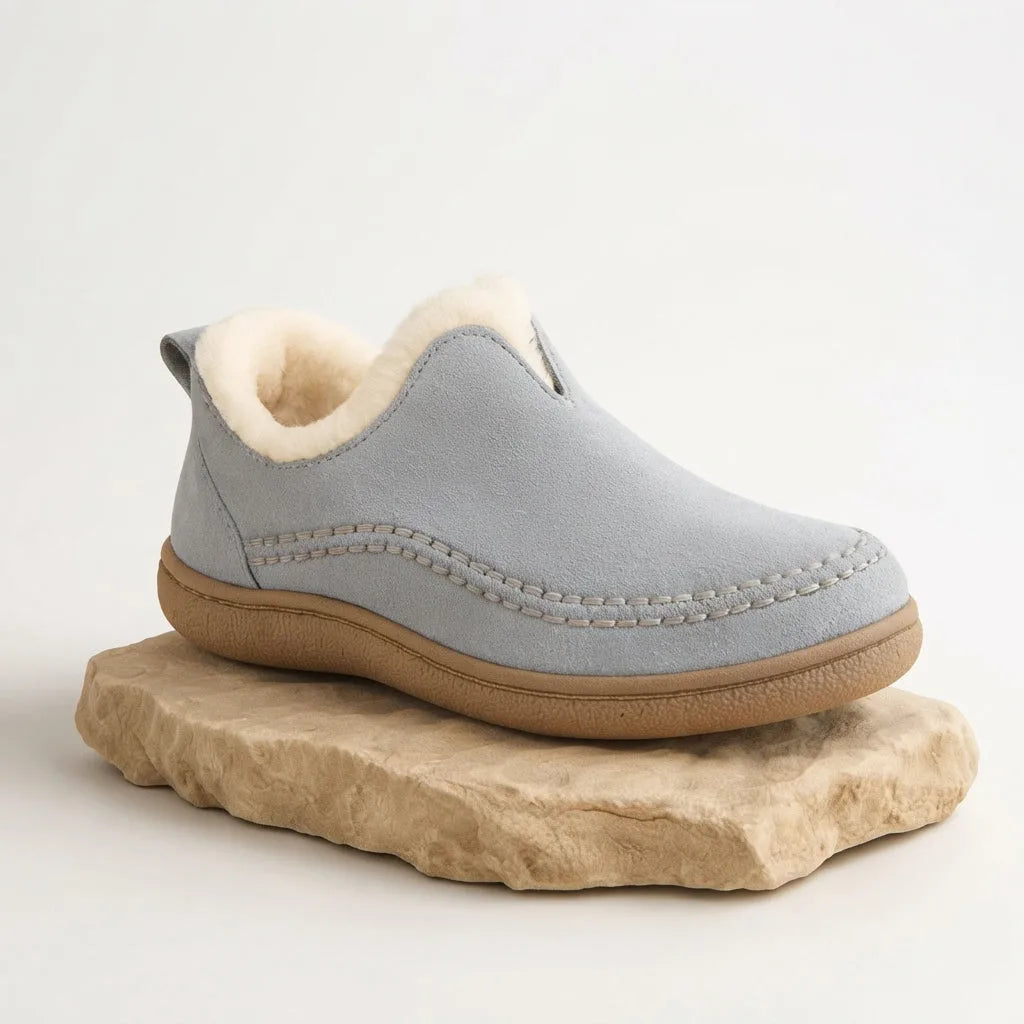 Side view of a pale grey suede slip-on boot with cream shearling trim, shown on a natural stone plinth