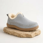 Side view of a pale grey suede slip-on boot with cream shearling trim, shown on a natural stone plinth