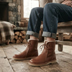 Model wearing brown leather slouch boots with turned-up jeans, seated in a cosy cabin setting