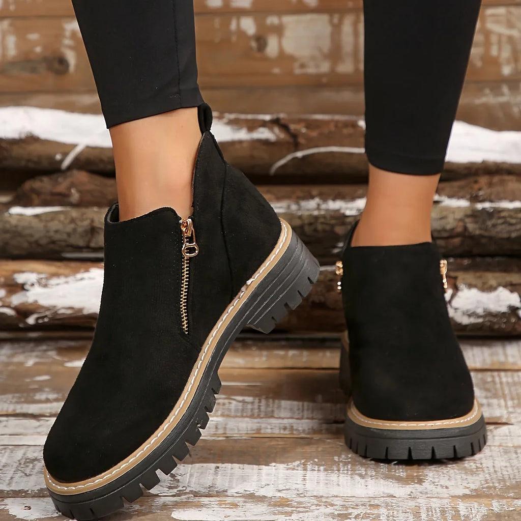 Black suede-style ankle boots with gold side zip and tan piping, worn crossed against a rustic wooden backdrop