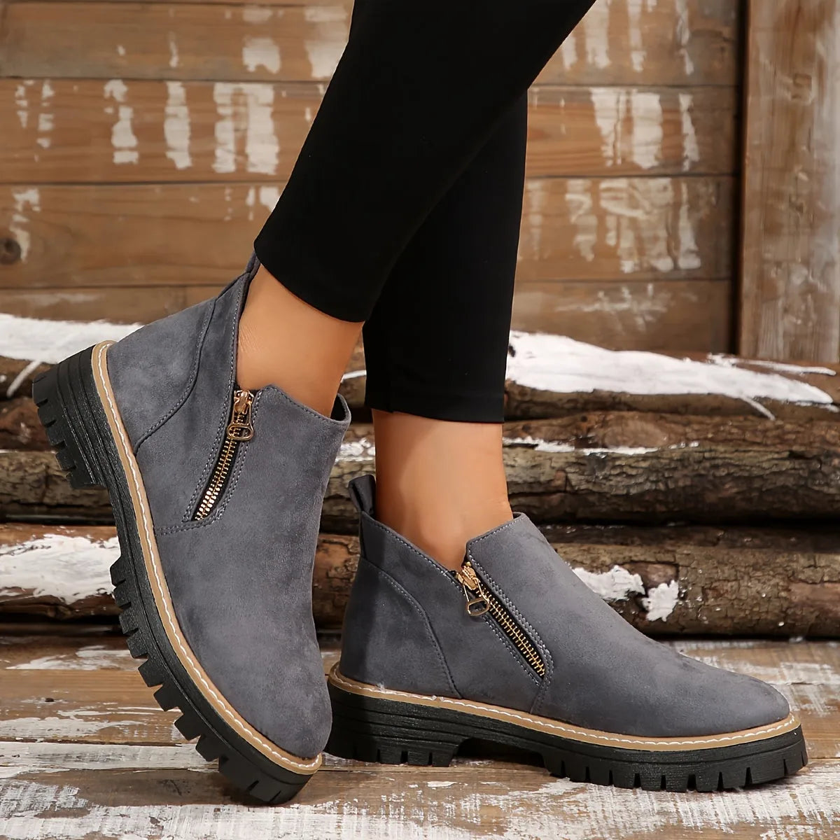 Grey suede-look ankle boots with gold zip fastening and tan stitched trim, worn on feet against wooden planks