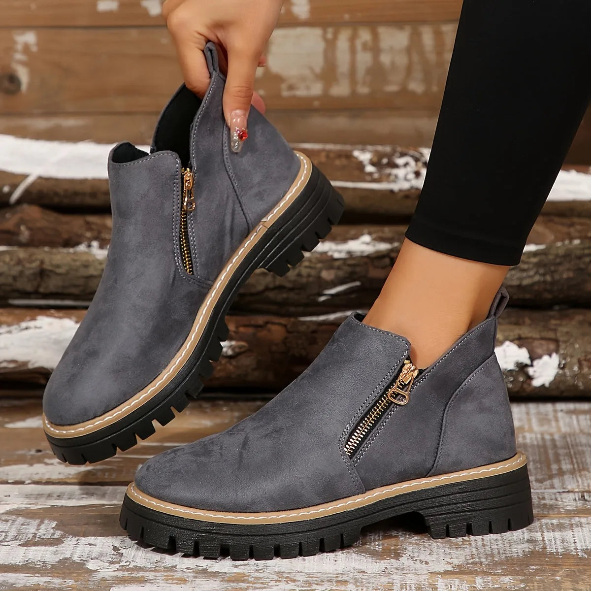 Pair of grey ankle boots held up by hand, showing the tan piping, gold zip pull and chunky tread sole
