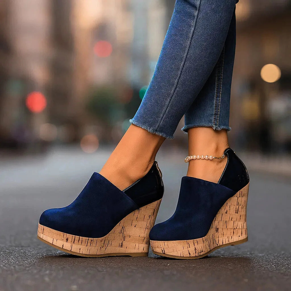 Close-up side view of navy suede wedge ankle boot with cork platform sole and patent heel trim, worn with cropped jeans and a pearl anklet