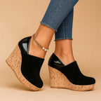 Studio side profile of black suede wedge ankle boots on a beige background, showing the patent trim and layered cork wedge