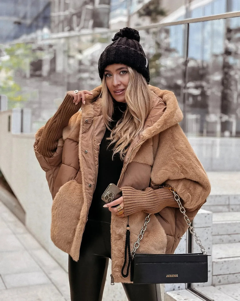 Woman wearing a camel oversized puffer coat with faux fur trim, hood up, carrying a black chain shoulder bag