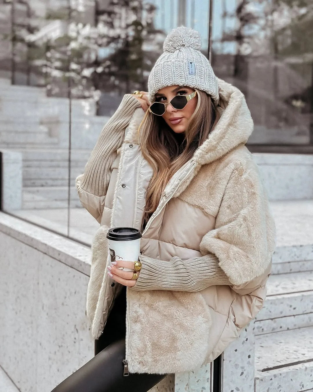 Beige oversized puffer coat with faux fur hood and panels, worn open over a black top with a pom pom beanie