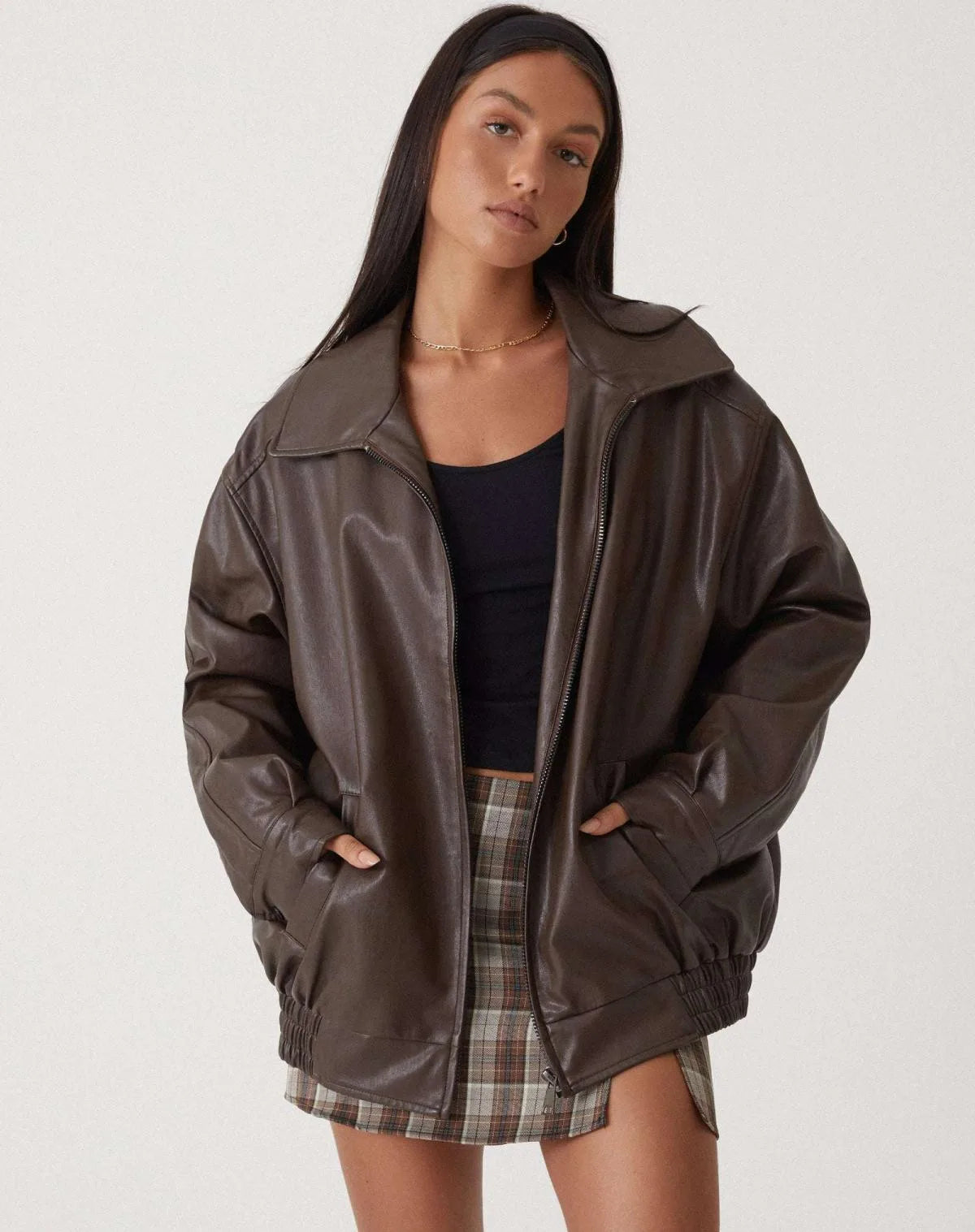 Model wears an oversized brown faux leather jacket open over a navy crop top and checked mini skirt