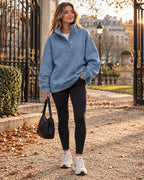 Woman wearing a mid-blue oversized henley sweatshirt with raised collar and dropped shoulders, paired with black leggings near an iron garden gate.
